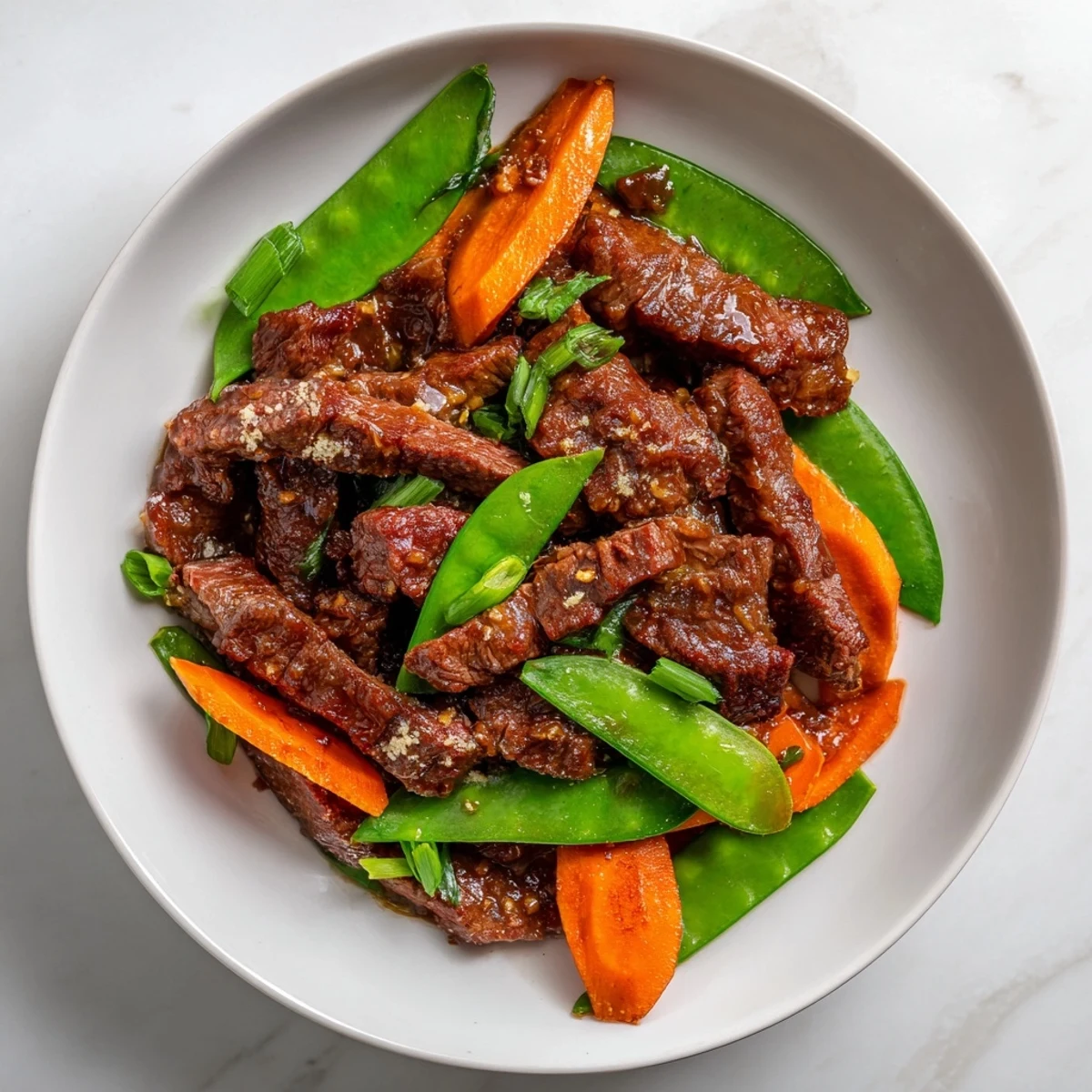 A steaming serving of beef stir fry with snow peas, carrots, and ginger over a bowl of fluffy white rice.