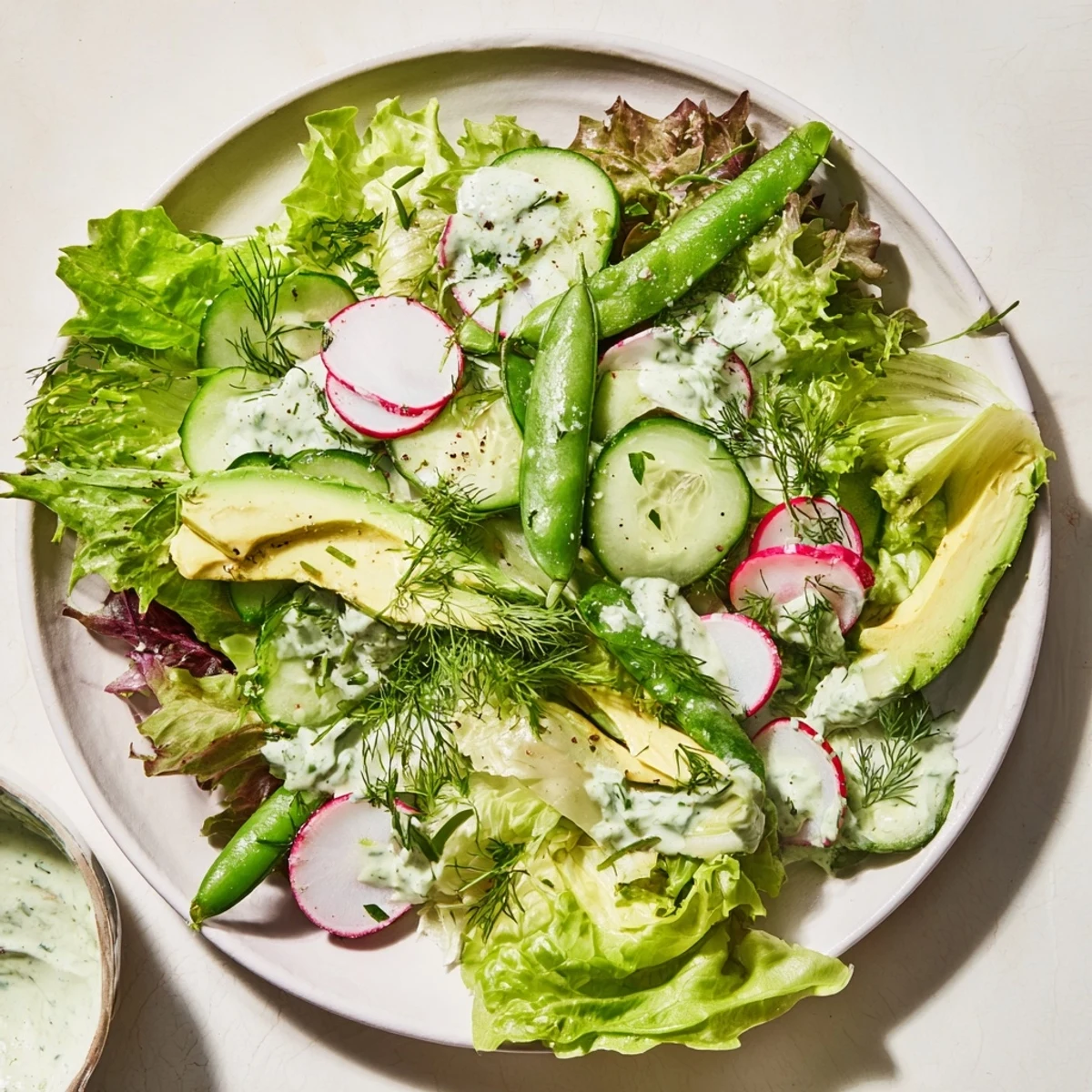 A vibrant bowl of Green Salad with Green Goddess Dressing, featuring crisp romaine, cucumber, avocado, and radishes glistening with herbaceous creamy dressing.  