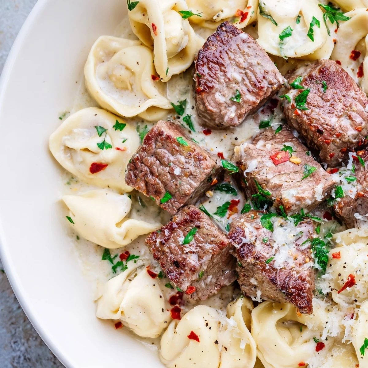 Freshly seared steak bites and tender cheese tortellini in a creamy garlic sauce, garnished with parsley. Garlic Steak Tortellini is served hot alongside a glass of red wine for a cozy dinner.