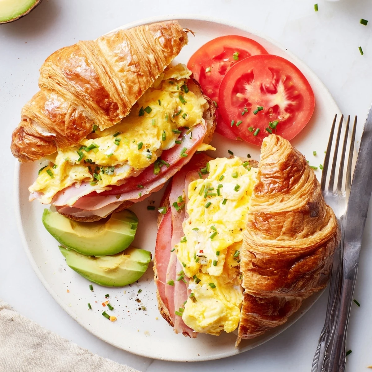 Flaky Croissant Breakfast Sandwich on a white plate, layered with ham and ripe avocado.