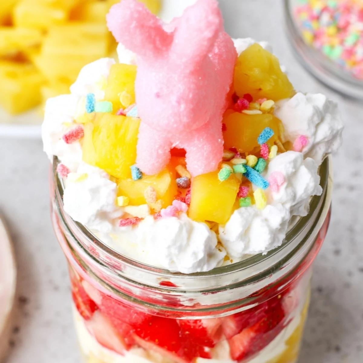 A close-up of the Jello Pie Easter Bunny Parfait In A Jar, showcasing vibrant layers of red strawberry and yellow lemon jello cubes, fresh diced fruit, and creamy filling topped with a white marshmallow bunny candy.