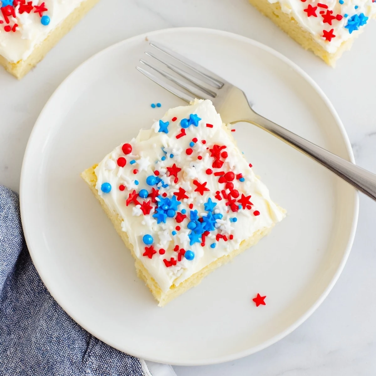Square sugar cookie bars with smooth vanilla frosting and scattered red white and blue sprinkles on a white serving plate
