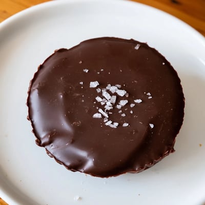 Paper liners cradle glossy chocolate cups topped with flaky sea salt, ready for a sweet snack.