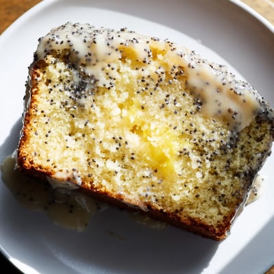 Thick slice of moist Lemon Poppy Seed Loaf showing crunchy seeds and tangy glaze on a plate.